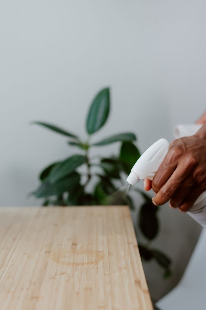 Office Cleaning A hand using a spray bottle to clean a wooden table indoors, with a plant in the background.